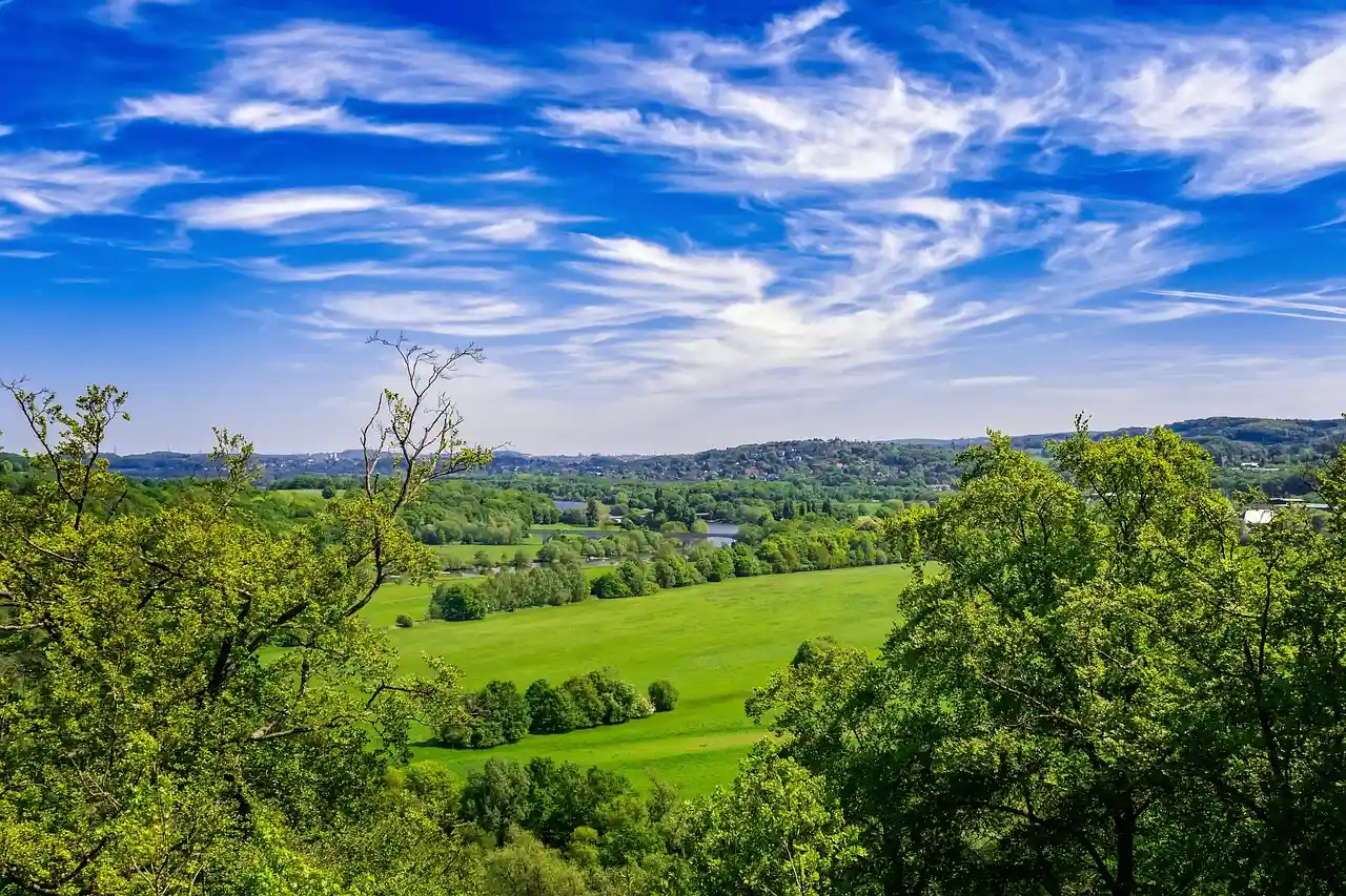 Blick von einer Anhöhe über grüne Wiesen und Baumgruppen, darüber blauer Himmel