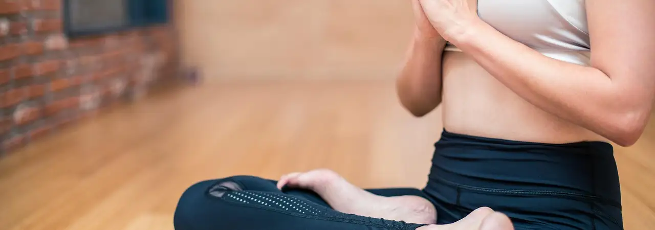 Person sitzt im Schneidersitz mit gefalteten Händen auf einer Yogamatte